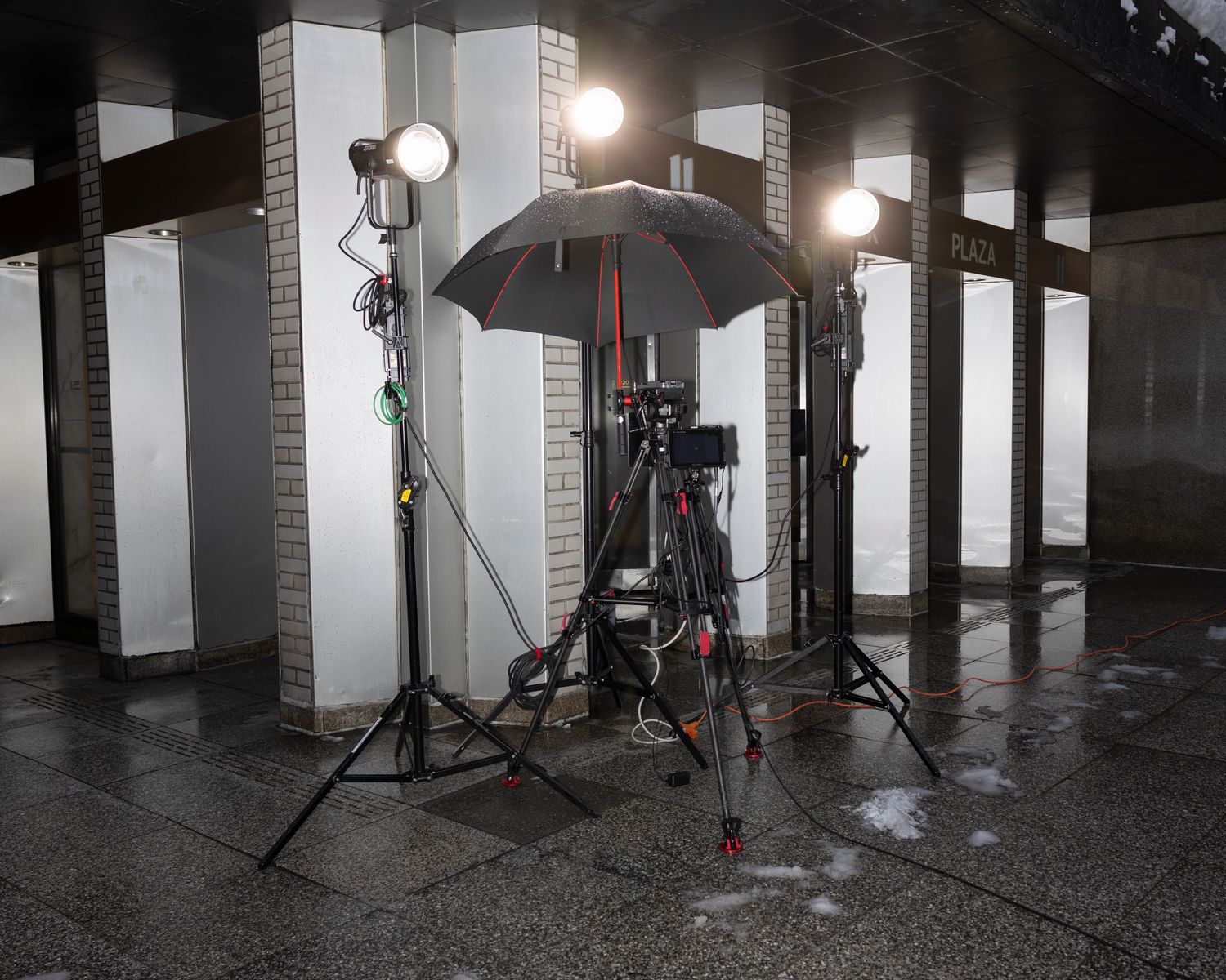 Professional film camera on a tripod between two production lights on a wet city plaza at night